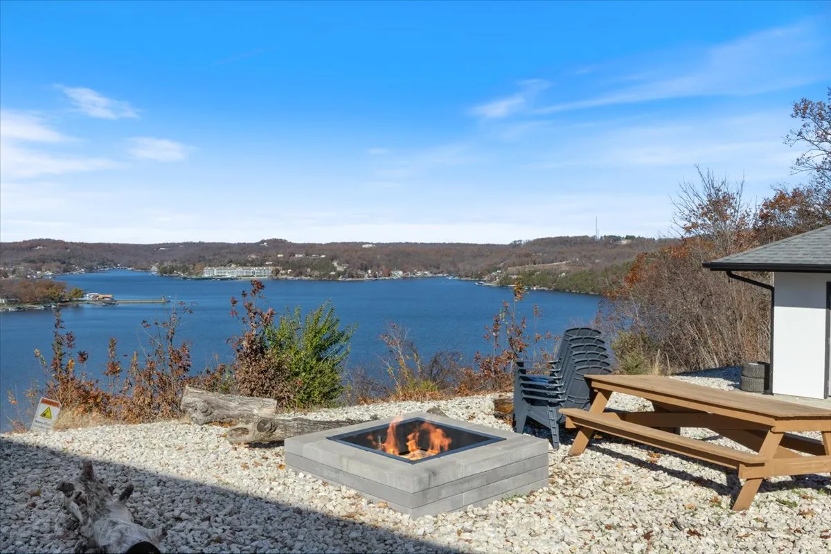 A cozy lakeside firepit area with inviting chairs, a picnic table, and direct views of the water. The space is set up for gatherings around the fire, casual picnics, and relaxing by the lake as the sun sets.