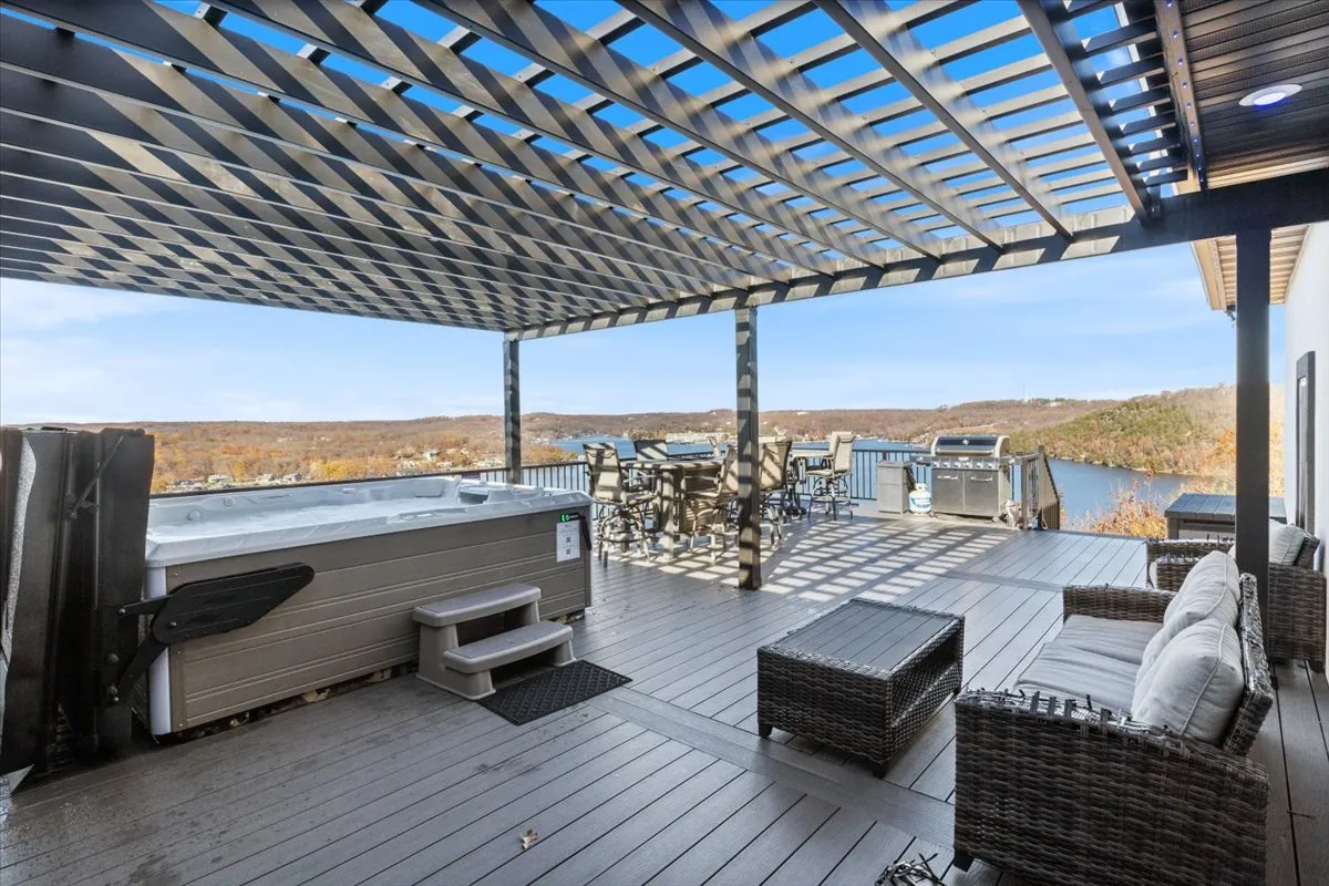 An inviting exterior scene featuring a spacious hot tub, outdoor dining set, lounge seating, and a scenic view of the lake. The atmosphere is relaxed and social, with clear sightlines to the water, ideal for group gatherings and sunset enjoyment.