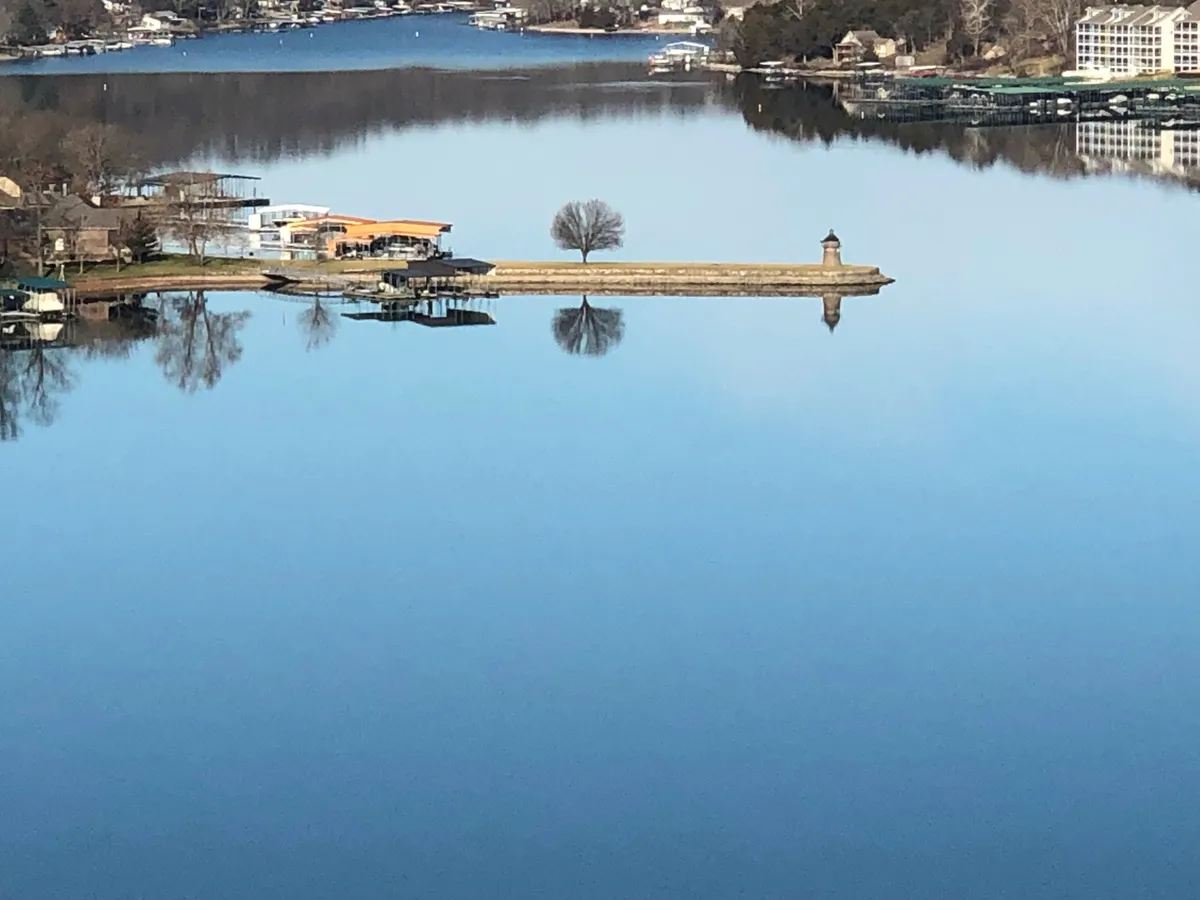 A tranquil lake scene with a calm water surface, a dock shaded by a tree, and homes dotting the distant shoreline. The peaceful vibe invites contemplation, boating, fishing, or simply enjoying the natural beauty at sunset.