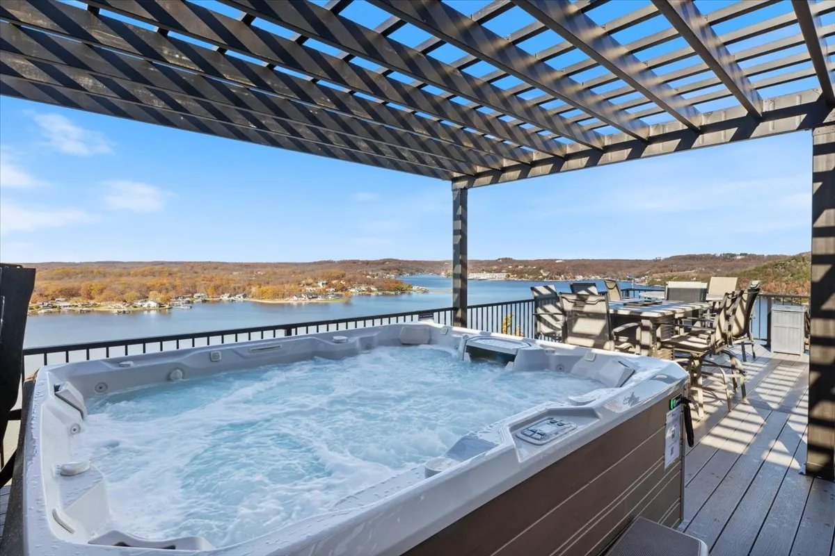 The image showcases a spacious deck overlooking the shimmering lake at sunset, featuring a bubbling hot tub, comfortable outdoor seating, and a dining area ideal for gatherings. The serene water view dominates the background, while the inviting hot tub and ample space for relaxation or entertaining are front and center.