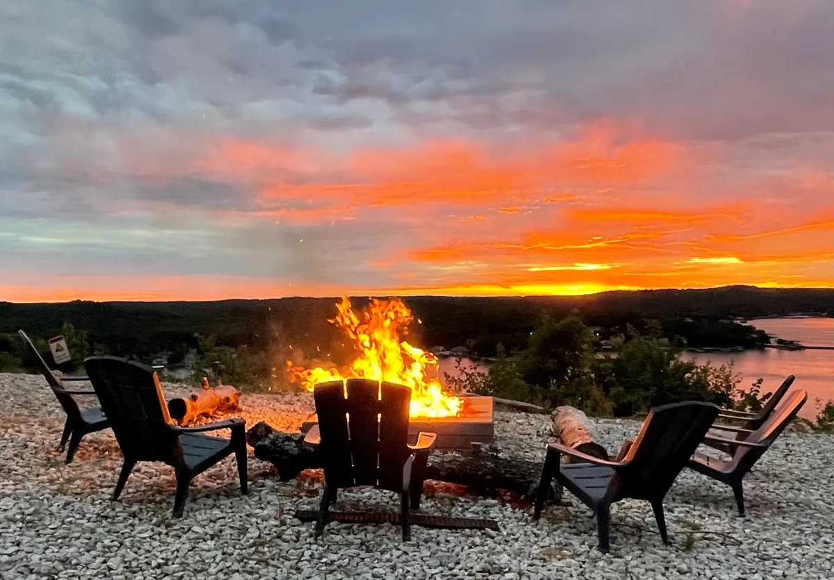 A cozy fire pit area surrounded by Adirondack chairs, set against a stunning sunset sky. The glow from the fire creates an inviting atmosphere for families or friends to gather, share stories, and enjoy the outdoors. The lake is visible in the distance, with the sky painted in warm hues.
