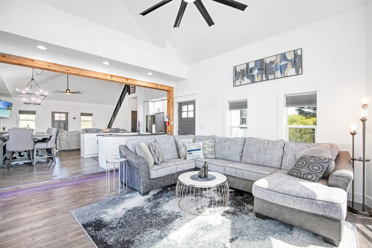 A stylish, modern living space featuring a large sectional sofa, a coffee table, prominent light fixtures, and an open view into a contemporary kitchen. Natural light pours in, and the configuration is perfect for socializing and entertaining.