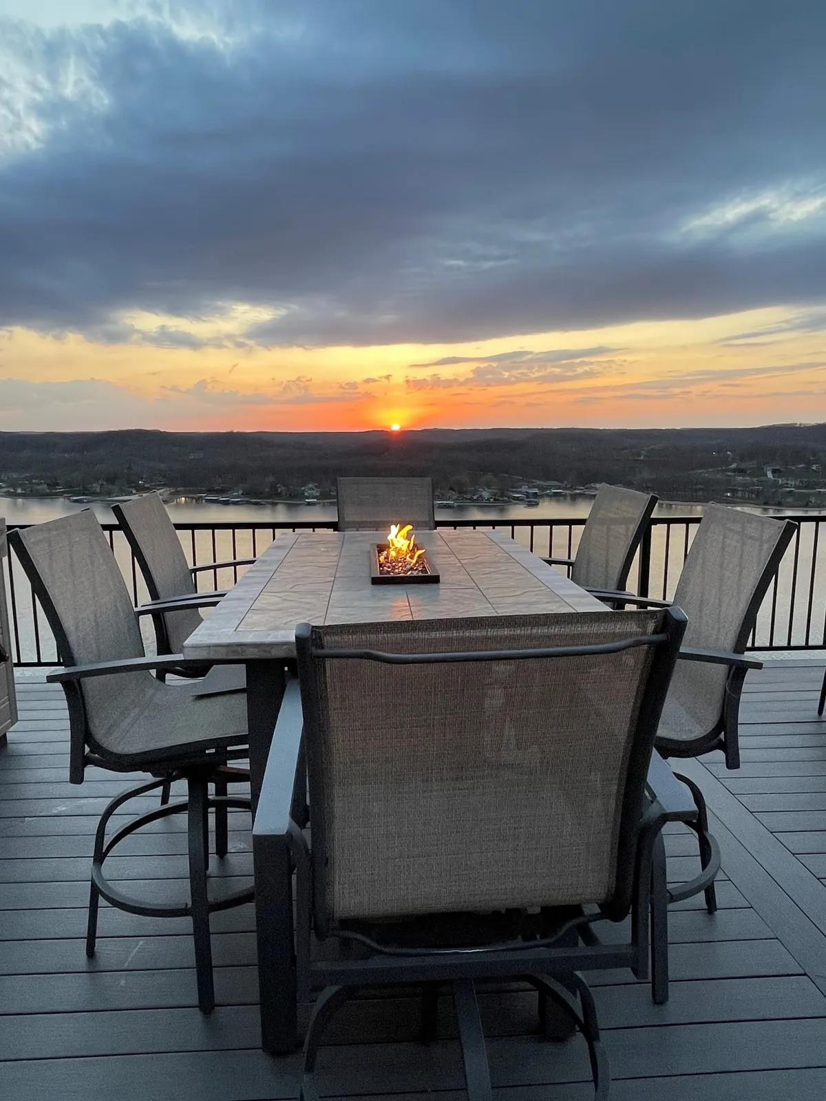 An inviting exterior scene with a dining table set near a glowing fire pit, all set against a dramatic sunset over the lake. The tranquil mood is underscored by the warm firelight, perfect for gathering with friends or family in the fresh air.