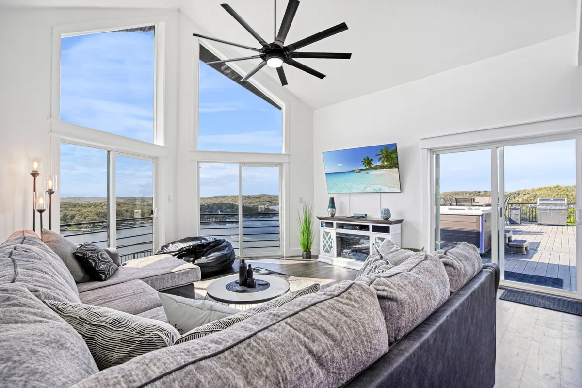 A comfortable sectional sofa anchors a spacious living room, set before large windows that frame a breathtaking lake view. Modern furnishings and a flat-screen TV (ideal for movie nights) round out the space, with serene lighting and décor emphasizing relaxation and togetherness.