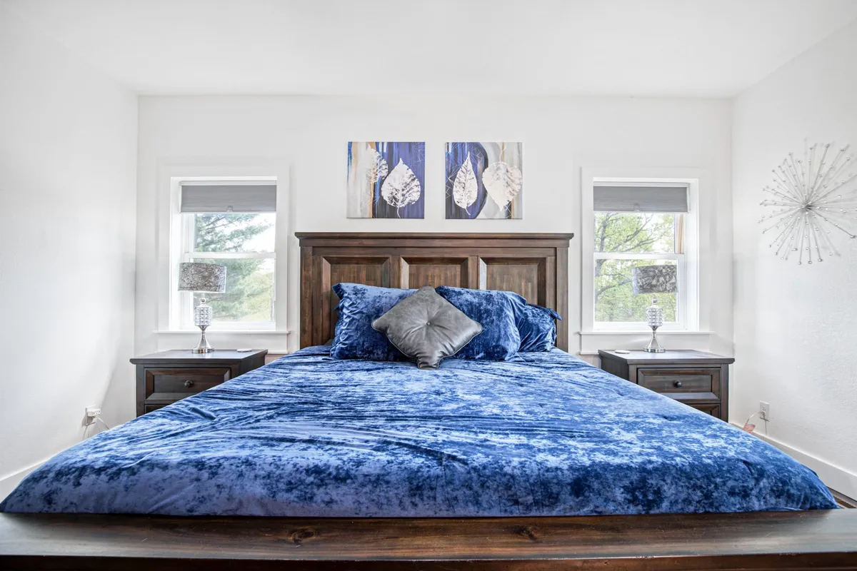 A serene bedroom featuring a large bed with plush blue velvet bedding, nightstands with softly glowing lamps, and abstract wall art. Gentle sunlight streams through the windows, creating a tranquil and cozy atmosphere ideal for unwinding.
