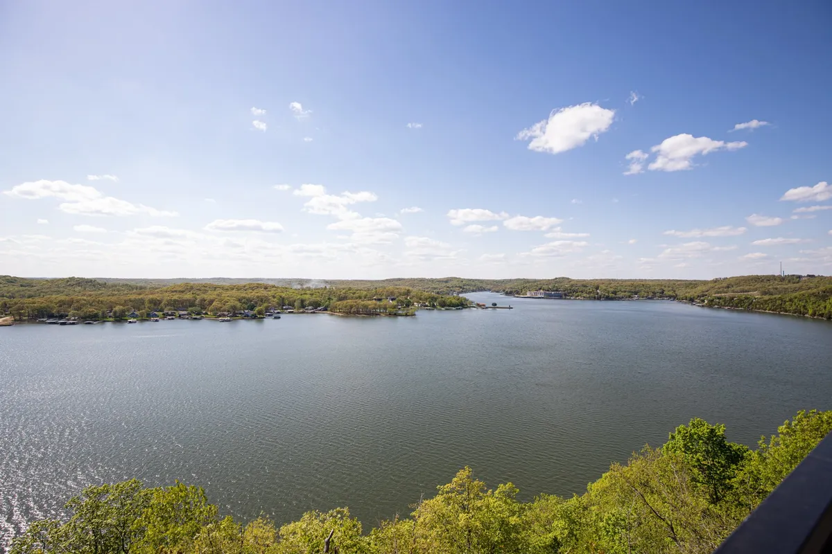 A sweeping view of the expansive lake surrounded by lush greenery and distant hills, with a cloud-filled sky creating an atmosphere of tranquility and natural beauty. The scenic vista invites exploration and highlights the outdoor opportunities available year-round.