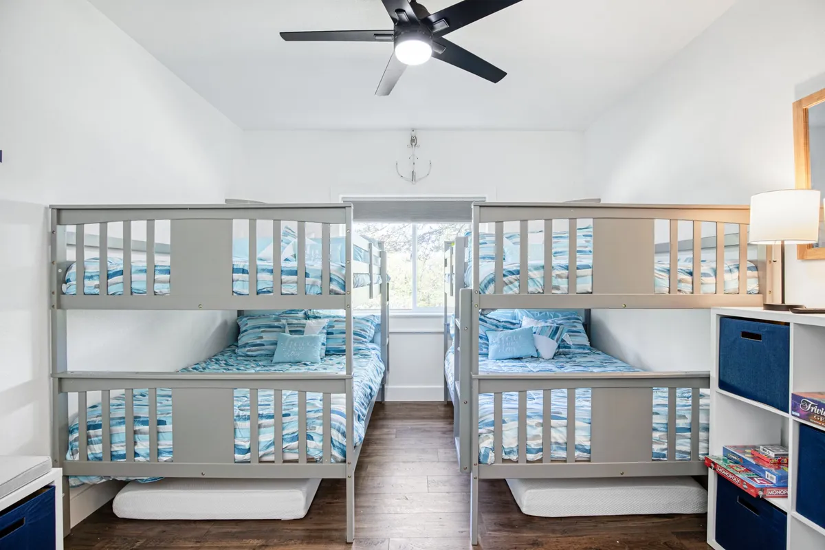 A playful and inviting bunk room designed for groups or families, with multiple bunk beds dressed in cheerful bedding, a window offering natural light, and a decorative nautical anchor on the wall. The space suggests a fun, social environment perfect for children or groups.