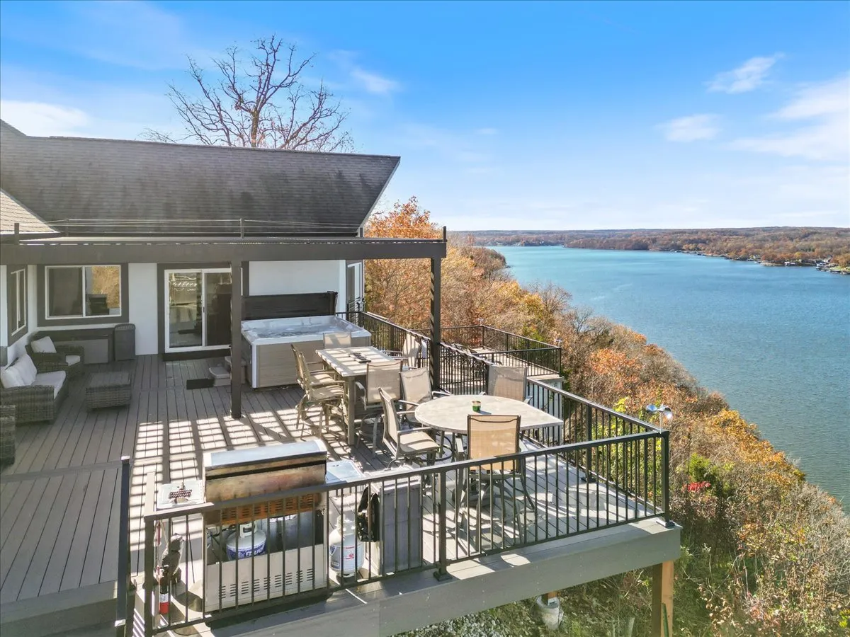 A spacious deck overlooking the water features a luxurious hot tub, a comfortable outdoor dining set, and serene river views. The space is designed for relaxation, with open skies and water providing a tranquil backdrop, perfect for enjoying both sunny days and cozy evenings.